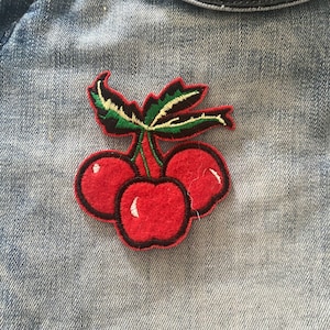 Może przedstawiać: Naszywka z haftowanych czerwonych wiśni z zielonymi liśćmi i łodygami. Naszywka znajduje się na niebieskim denimie.