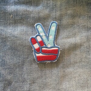 Puede incluir: Un parche con forma de signo de paz con estrellas y rayas rojas, blancas y azules. El parche está hecho de fieltro y tiene un borde cosido.