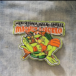 Peut inclure: Patch brodé vert, orange et jaune représentant un personnage de la Tortue Ninja avec le texte "HEROES IN A HALF-SHELL! MICHELANGELO".