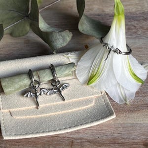Può includere: Set di gioielli a tema drago color argento. Il set include un anello e un paio di orecchini a cerchio, ciascuno con un design a drago. Gli orecchini e l'anello sono esposti con un fiore bianco e un piccolo astuccio.