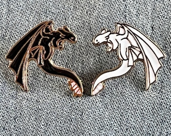 May include: Two enamel pins depicting dragons, one black and one white, facing each other. The dragons are stylized and have wings and tails. The pins are a popular collectible for fans of fantasy and mythical creatures.
