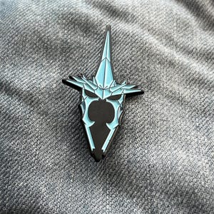 Puede incluir: Un pin de esmalte azul y negro con un diseño estilizado y geométrico de la cabeza de una criatura con dientes afilados y una corona puntiaguda.