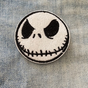 May include: White and black embroidered patch of a cartoon character's face with a stitched smile. The character has black eyes and a white face.