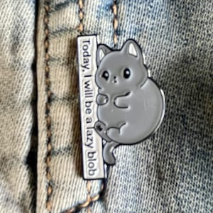 Op de afbeelding: Een grijze emaille pin met een cartoonkat met de tekst "Today, I will be a lazy blob" op een witte achtergrond.