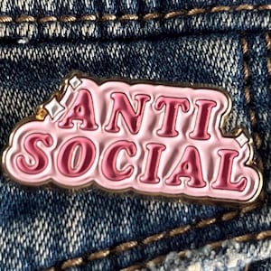 Könnte beinhalten: Ein rosa-goldener Emaille-Pin mit den Worten "ANTI SOCIAL" in einer Bubble-Letter-Schrift. Der Pin ist vor einem Jeans-Hintergrund platziert.