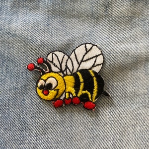 Puede incluir: Un parche bordado de abeja con rayas amarillas y negras, patas rojas y un ala blanca y negra. La abeja sonríe y tiene ojos grandes. La abeja está sobre un fondo de mezclilla azul.
