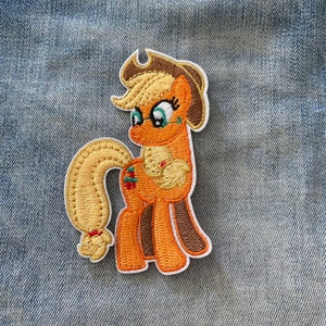 Op de afbeelding: Een geborduurde patch van een cartoonpony met een bruine hoed, blond haar en een bril. Het pony is oranje en bruin met een gele staart. De patch is op een blauwe denim achtergrond.