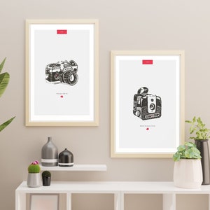 Puede incluir: Dos ilustraciones en blanco y negro de cámaras vintage enmarcadas en una pared blanca. Las ilustraciones están sobre un fondo blanco con texto rojo. Los marcos son de madera clara. Las ilustraciones están colgadas en una pared blanca sobre un estante blanco con un jarrón, dos plantas pequeñas y una maceta blanca.