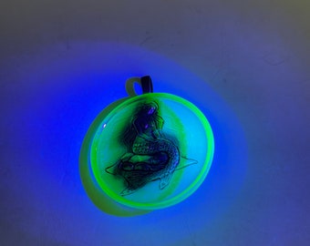 May include: A round, glowing pendant with a mermaid design. The pendant has a black mermaid silhouette inside a clear, circular frame with a neon green edge. A black loop is attached to the top.