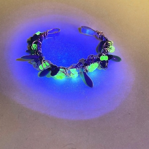 May include: A handmade bracelet with a wire-wrapped design. The bracelet features green and black beads and leaf-shaped accents. The beads and accents glow with a bright green color. The bracelet is set against a blue and white background.