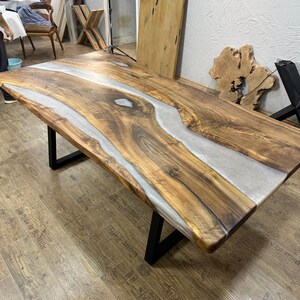以下が含まれることがあります： ライブエッジデザインの大きな長方形の木製テーブル。テーブルの中央には、薄いグレーのエポキシ樹脂の川があります。木材は自然な温かみのある茶色で、脚は黒い金属です。