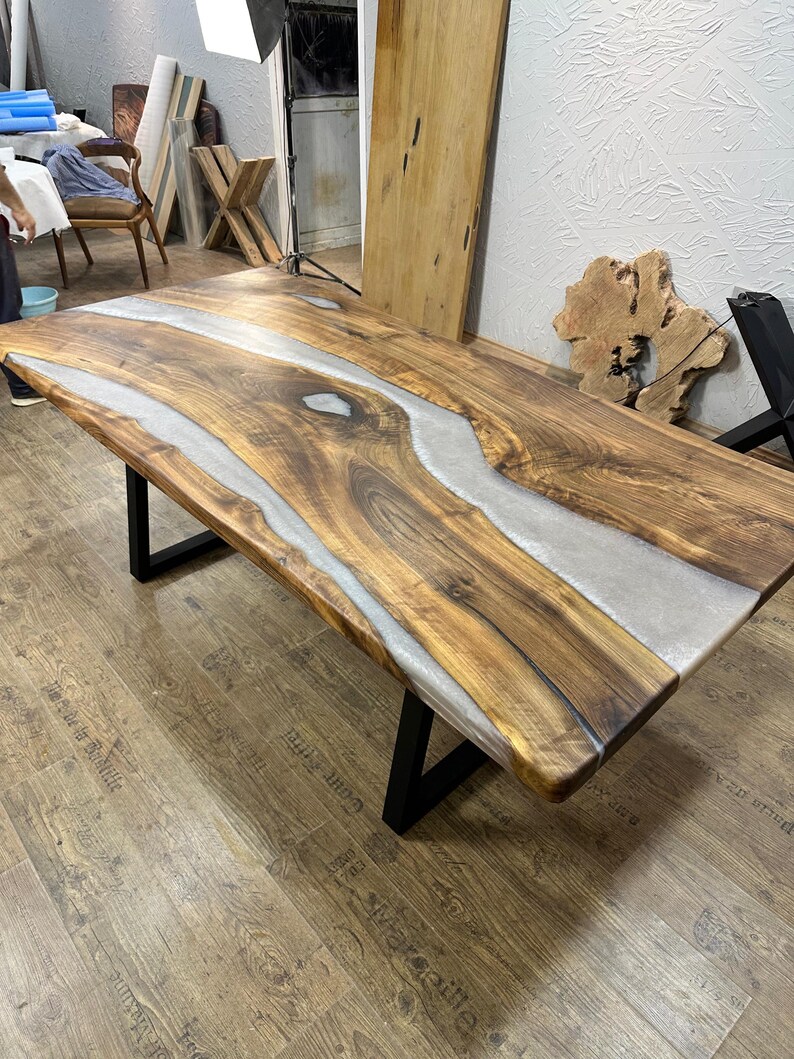 以下が含まれることがあります： ライブエッジデザインの大きな長方形の木製テーブル。テーブルの中央には、薄いグレーのエポキシ樹脂の川があります。木材は自然な温かみのある茶色で、脚は黒い金属です。