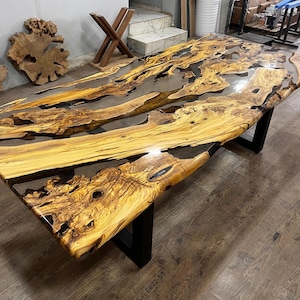 May include: A large, rustic wooden table with a live edge top and black metal legs. The table top is made of wood with a natural grain and is covered in a clear epoxy resin. The resin has a flowing, river-like pattern and creates a unique and beautiful look.