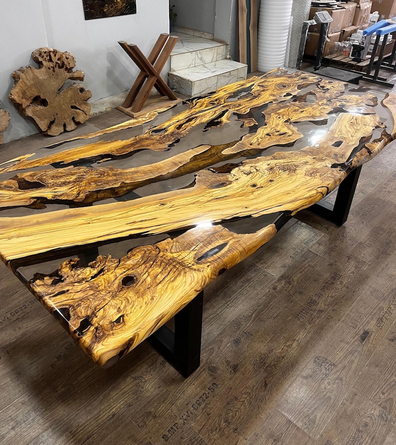 May include: A large, rustic wooden table with a live edge top and black metal legs. The table top is made of wood with a natural grain and is covered in a clear epoxy resin. The resin has a flowing, river-like pattern and creates a unique and beautiful look.