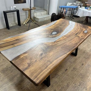 以下が含まれることがあります： ライブエッジデザインの大きな長方形の木製テーブル。テーブルの中央には銀色のエポキシ樹脂の川があります。木製の表面は温かみのある茶色で、テーブルは黒い金属製の脚で支えられています。