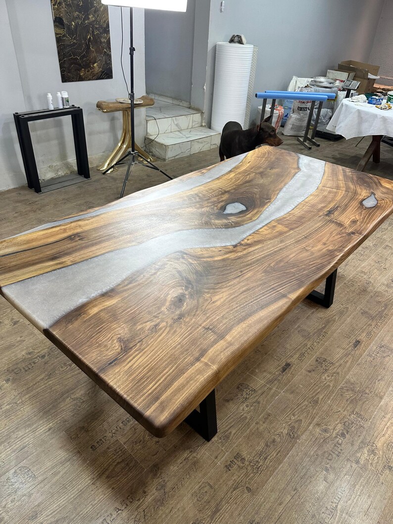 以下が含まれることがあります： ライブエッジデザインの大きな長方形の木製テーブル。テーブルの中央には銀色のエポキシ樹脂の川があります。木製の表面は温かみのある茶色で、テーブルは黒い金属製の脚で支えられています。