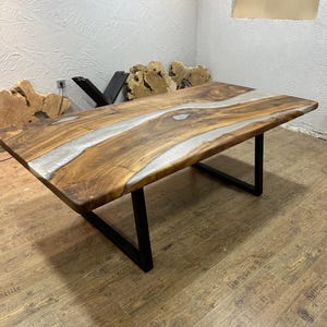 以下が含まれることがあります： ライブエッジデザインと透明なエポキシ樹脂の川がある長方形の木製テーブル。テーブルは天然の木目仕上げで、黒い金属製の脚で支えられています。テーブルの長さは約183cmです。