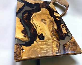Op de afbeelding: Een rechthoekige salontafel met een uniek blad van hout en epoxyhars. Het blad heeft een natuurlijk houtnerfpatroon met zwart en grijs epoxyhars dat erdoorheen stroomt. De tafel heeft een eenvoudige zwarte metalen basis.