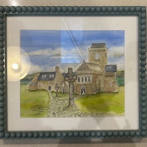 May include: A watercolour painting of a church with a cross in front of it. The church is made of stone and has a tall steeple. The cross is made of wood and has a simple design. The painting is framed in a blue frame with a beaded edge. The text "Inna Athene and In Memories of my love, you are always in my heart.  D.B.A.F." is written in the bottom left corner of the painting.
