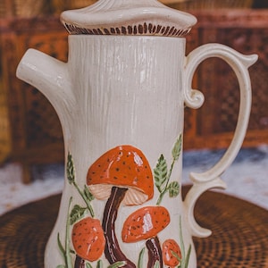 May include: Cream ceramic teapot with a lid and handle, decorated with orange mushroom illustrations and green foliage. The teapot has a textured surface and a dark brown mushroom-shaped knob on the lid. A decorative kitchenware item.