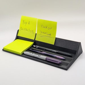 Puede incluir: Un organizador de escritorio de plástico negro con tres ranuras para bolígrafos y dos ranuras para notas adhesivas. Las notas adhesivas tienen mensajes escritos a mano: "Comprar una bandeja de notas Post-it XL" y "Echa un vistazo a Streno Goods".