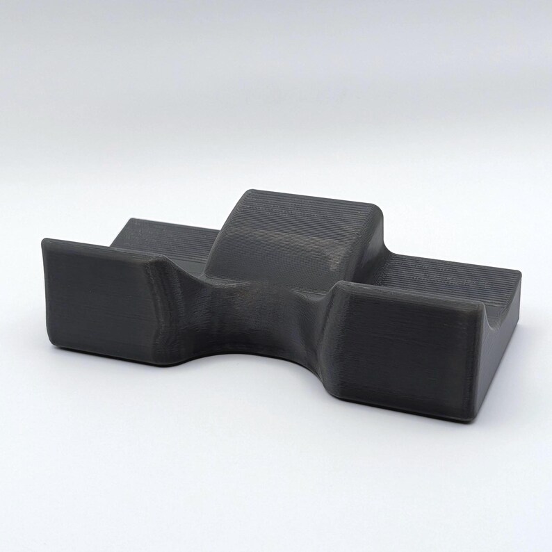 May include: A dark gray, 3D-printed object with a unique shape. It features a central raised section flanked by two lower, rectangular platforms. The object has a smooth, matte finish and appears to be made of a solid material.