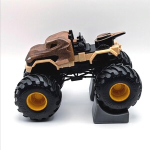 May include: A brown and tan monster truck toy with large black tires and yellow rims. The truck has a detailed body design and is set on a gray stand. The toy truck is designed for play.