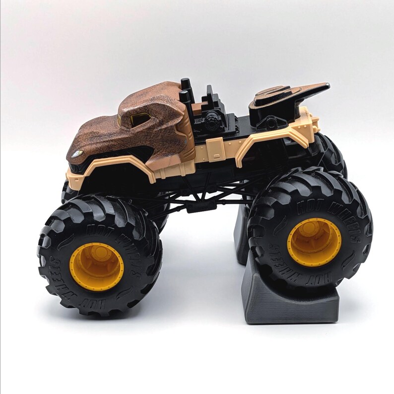 May include: A brown and tan monster truck toy with large black tires and yellow rims. The truck has a detailed body design and is set on a gray stand. The toy truck is designed for play.