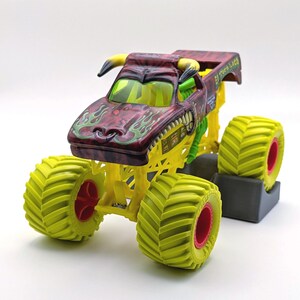 May include: A toy monster truck with a maroon body, yellow chassis, and large lime green tires with red rims. The truck features a bull-like design with yellow horns and the text "El Toro Loco".