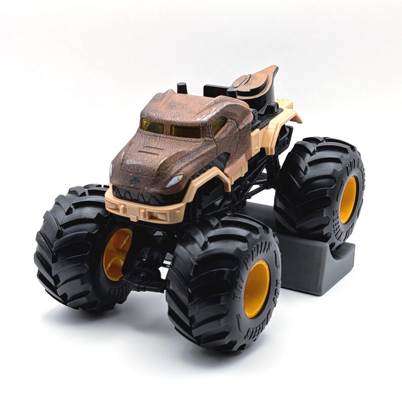 May include: A brown and tan monster truck toy with large black tires and yellow rims. The truck has a detailed design on the cab and body, and is set against a white background.