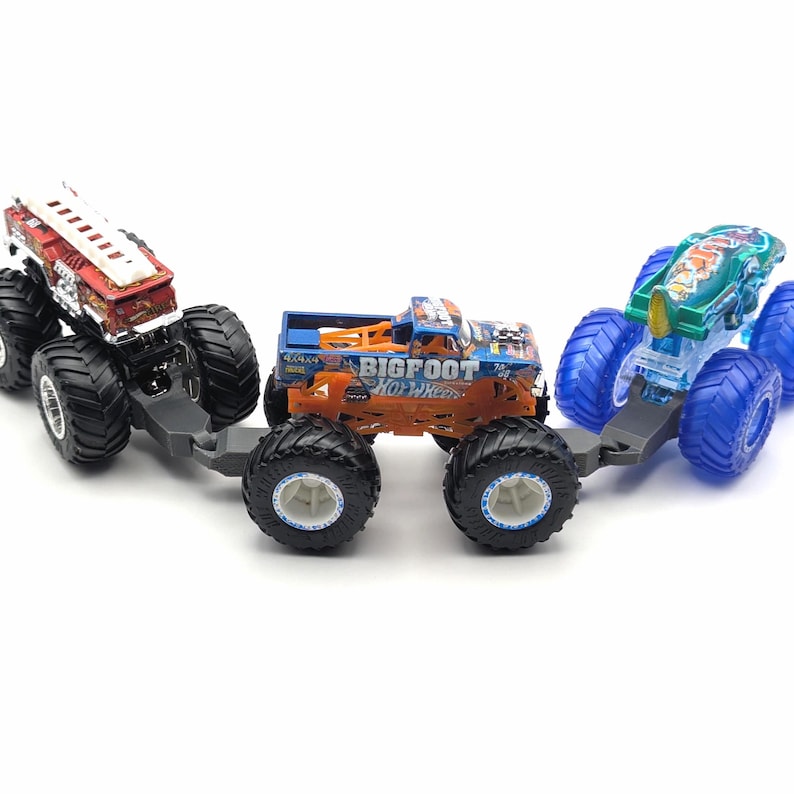 May include: Three toy monster trucks, one red and white fire truck, one orange and blue monster truck with "BIGFOOT" and "HO WHEELS" printed on the side, and one blue and green monster truck. The trucks are connected together.