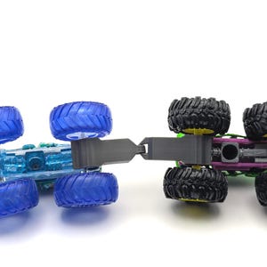 May include: Two toy monster trucks connected together. The front truck is blue with clear plastic body and four blue wheels. The rear truck is green with black wheels and a purple chassis.