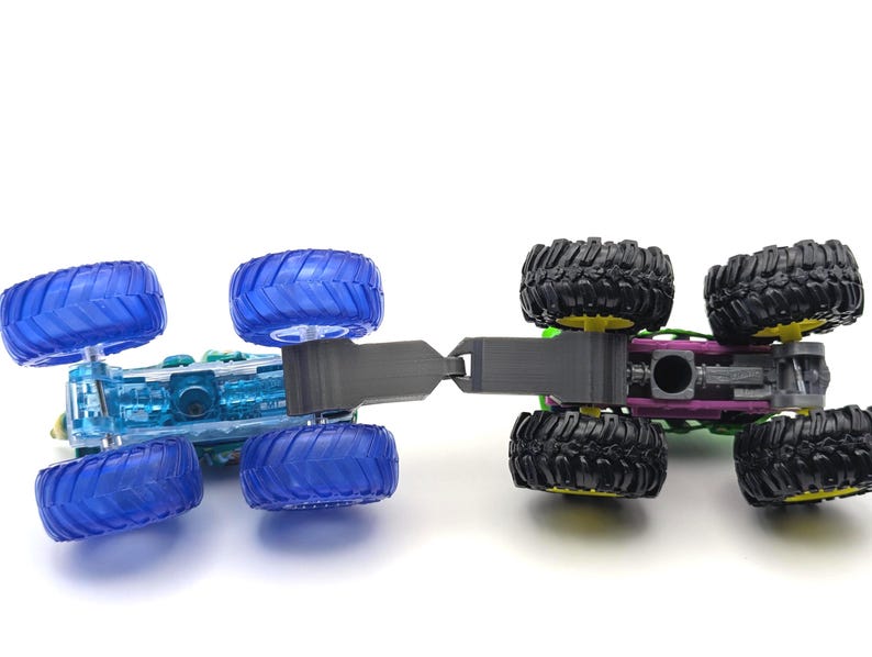May include: Two toy monster trucks connected together. The front truck is blue with clear plastic body and four blue wheels. The rear truck is green with black wheels and a purple chassis.