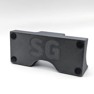 May include: A dark gray, rectangular object with rounded edges and a curved cutout. The letters "SG" are embossed on the front. Four small, black circular pads are affixed to the bottom of the object.