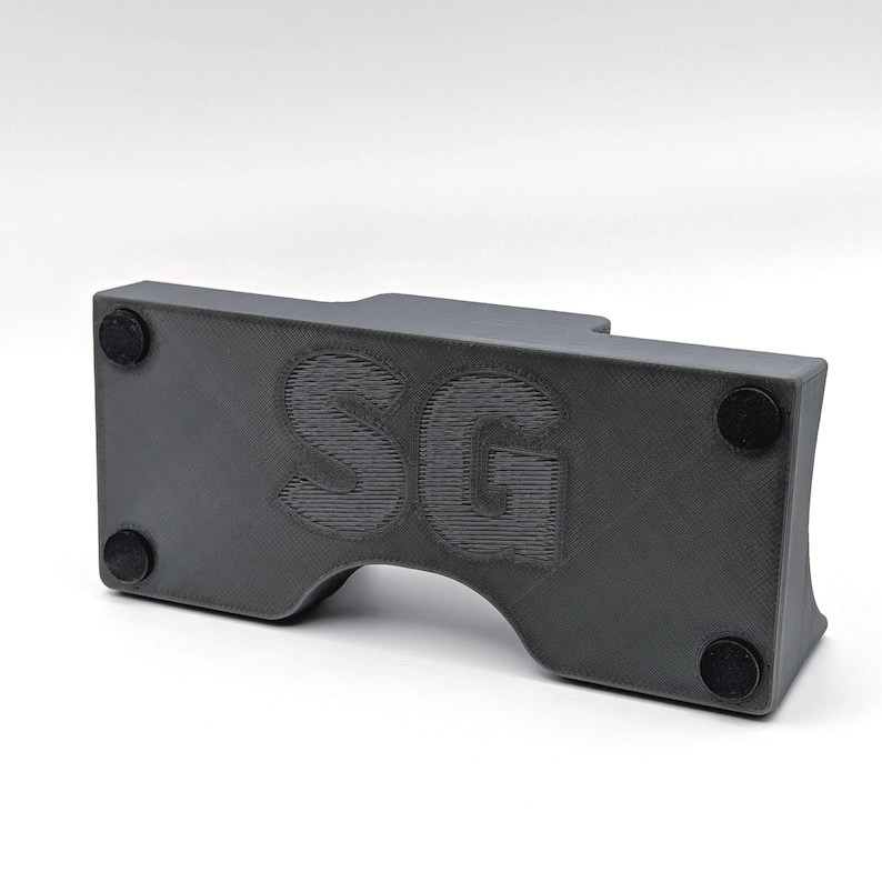 May include: A dark gray, rectangular object with rounded edges and a curved cutout. The letters "SG" are embossed on the front. Four small, black circular pads are affixed to the bottom of the object.