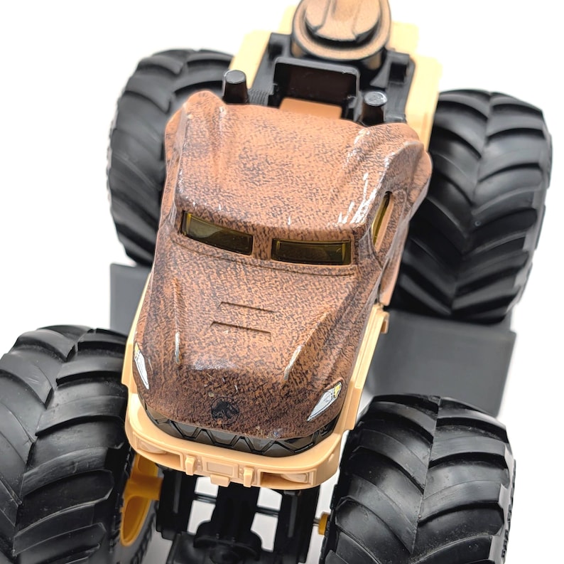 May include: A brown and tan monster truck toy with large black tires. The truck features a textured brown body, yellow tinted windows, and a tan chassis. The tires are thick and have a deep tread.