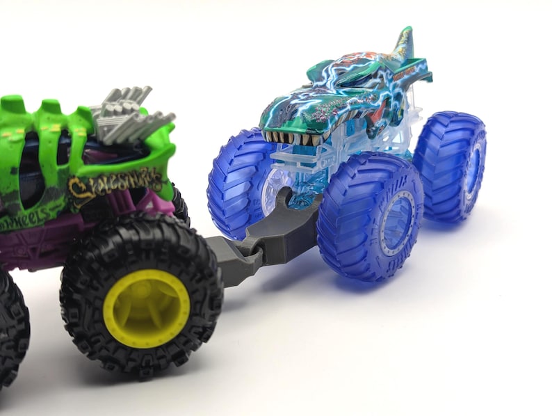 May include: Two toy monster trucks, one green and one blue, with large wheels. The green truck has a skull design and the text "Skeletal Wheels". The blue truck has a shark design and is translucent.