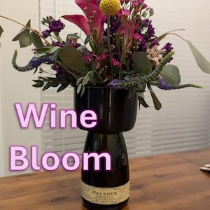 May include: A bouquet of purple, pink, and yellow flowers in a black vase made from a recycled wine bottle. The bottle is labeled "Paladin Prosecco". The text "Wine Bloom" is superimposed on the image.