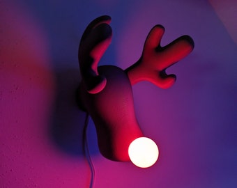 May include: A white light bulb illuminates the nose of a 3D printed reindeer head lamp. The reindeer is a dark red color and is mounted on a wall. The wall is painted a dark purple color.