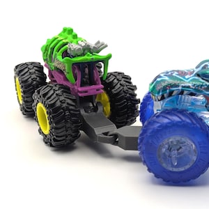May include: Two monster trucks, one green and one blue, are connected together. The green monster truck has a skeletal design and is pulling the blue monster truck. Both trucks have large wheels.