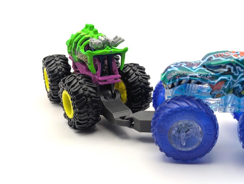 May include: Two monster trucks, one green and one blue, are connected together. The green monster truck has a skeletal design and is pulling the blue monster truck. Both trucks have large wheels.