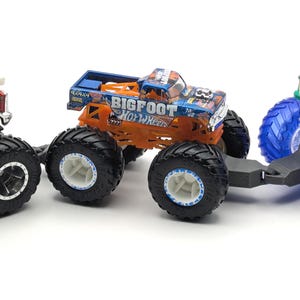 May include: Three toy monster trucks, one red and white, one orange and black with "BIGFOOT HOT WHEELS" on the side, and one blue and green. The orange and black truck is pulling the blue and green truck with a black tow hitch.