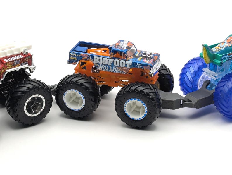 May include: Three toy monster trucks, one red and white, one orange and black with "BIGFOOT HOT WHEELS" on the side, and one blue and green. The orange and black truck is pulling the blue and green truck with a black tow hitch.