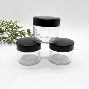 May include: Three clear plastic jars with black screw-on lids are stacked on a white surface. The jars are empty and cylindrical, with a wide opening. The lids are smooth and opaque, contrasting with the transparent bodies of the jars.