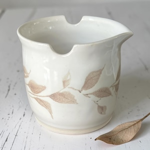 May include: A white ceramic creamer with a spout and a handle-less design. The creamer features a brown leaf pattern around the body. The creamer is sitting on a white wooden surface.