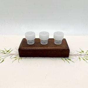 May include: Three small, clear plastic vials with white screw-on lids are arranged on a dark brown wooden block. The vials are evenly spaced and appear to be empty. The block sits on a decorative surface with a floral design.