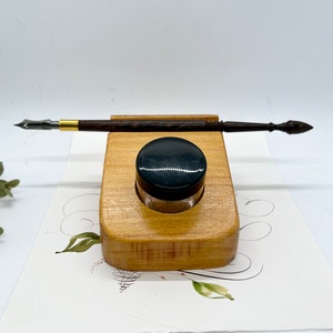 May include: A wooden pen holder with a dip pen and inkwell. The pen is dark brown with a gold-colored metal band. The inkwell is clear glass with a black lid. The holder is light brown and sits on a piece of paper with floral designs.
