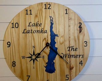 May include: A wooden clock with a blue lake outline in the center. The clock face has Roman numerals and a compass rose. The text "Lake Latonka" and "The Wimers" is printed on the clock face.