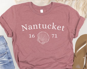 Puede incluir: Una camiseta color malva con texto blanco que dice "Nantucket 16 71" y un gráfico de concha blanca.