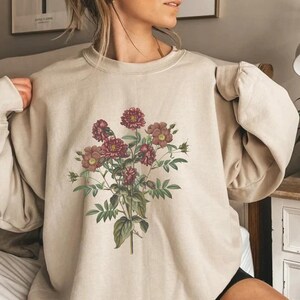 May include: A beige sweatshirt with a floral print featuring pink and red flowers. The flowers are arranged in a bouquet with green leaves and stems.